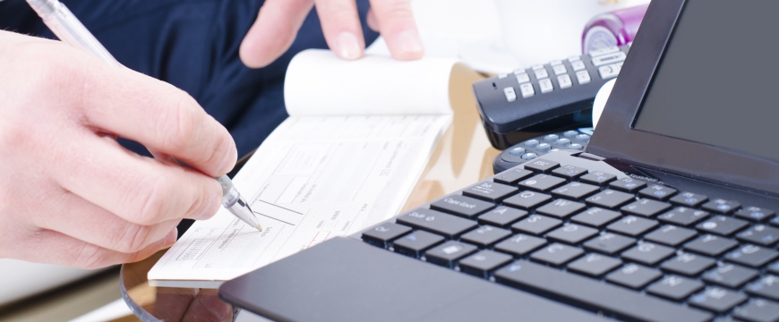 Business Checking, image of man signing checks with computer and calculator on desk