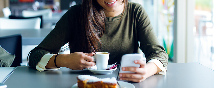 Mobile Banking - smiling girl seated at table with coffee cup in hand and mobile phone in other